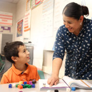 A teacher looks over the shoulder of a young boy in an orange shirt while he reads a page