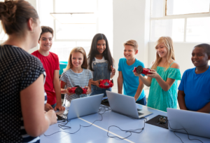 A group of students, both boys and girls, are gathered around a table with laptops, AI tools, and small robotic devices. They are smiling and appear to be engaged in a STEM or robotics activity. An instructor is standing in front of them, leading the session in a bright, well-lit classroom.