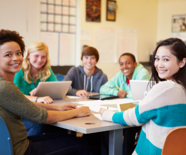 A group of students sitting around a table in a classroom, smiling and engaging with each other through SEL strategies. Some have laptops and notebooks in front of them, and the background shows a bulletin board and classroom materials.